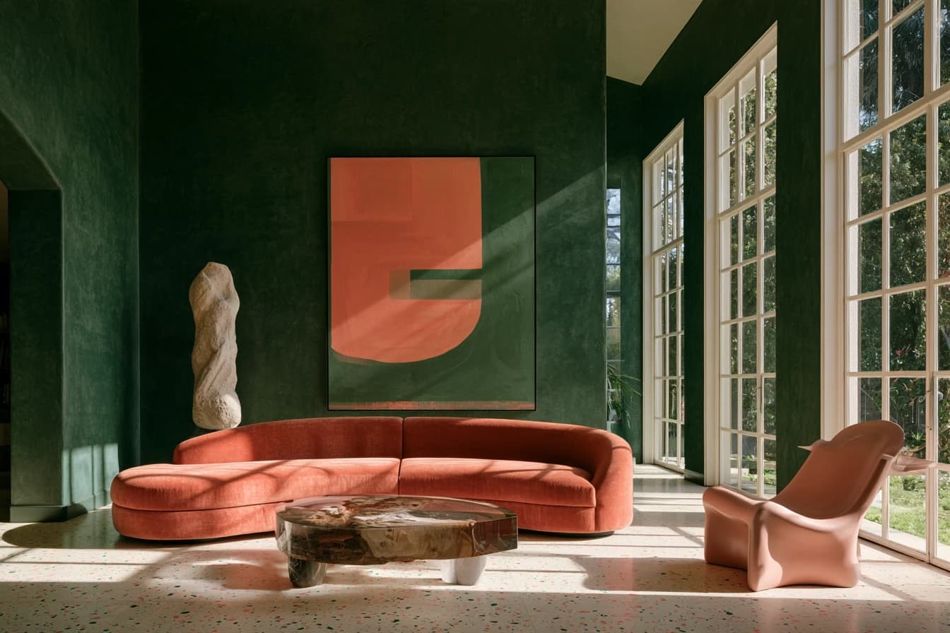 Living room with deep green plaster walls, a curved coral velvet sofa, a large coral and green painting, a stone sculpture and a terrazzo floor in raking sunlight